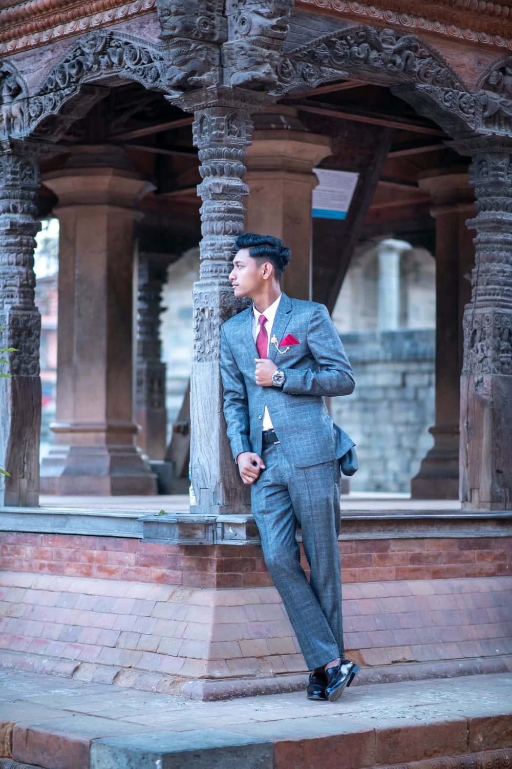 Deepak in a grey suit and red tie leaning against a carved wooden pillar of a traditional pavilion