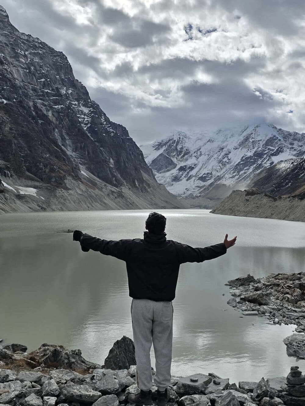 Deepak with arms outstretched facing a glacial lake below snowy peaks