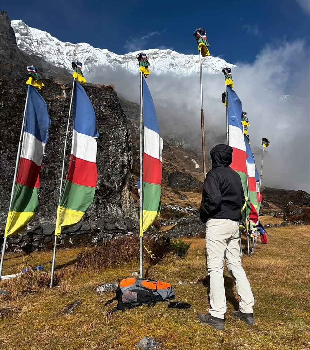 Deepak in a black hooded jacket facing a row of tall prayer flags below a snowy Himalayan peak